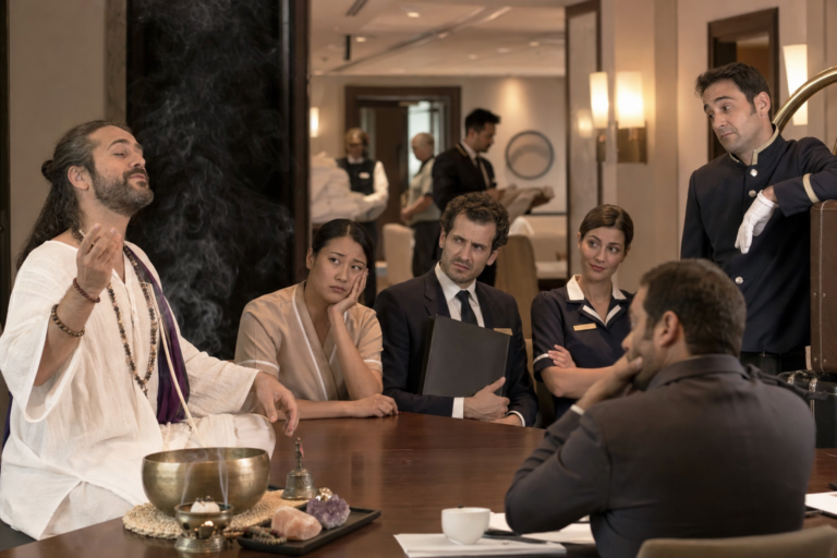 Falso gurú espiritual dando una charla ante un equipo hotelero visiblemente escéptico mientras la operación continúa al fondo